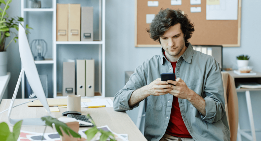 Young professional reviewing social media content on a smartphone at his desk, representing online screening during the hiring process.