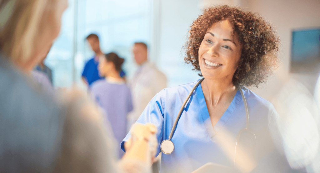 Smiling healthcare worker in blue scrubs with a stethoscope shakes hands with a patient in a bright medical office.